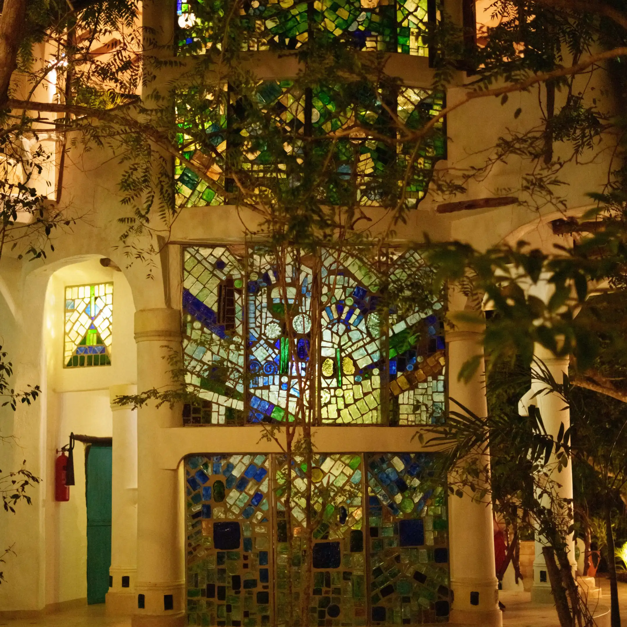 Interior courtyard wall covered with green, blue and yellow stained-glass mosaic panels, flanked by white columns and leafy indoor trees, softly lit at dusk.