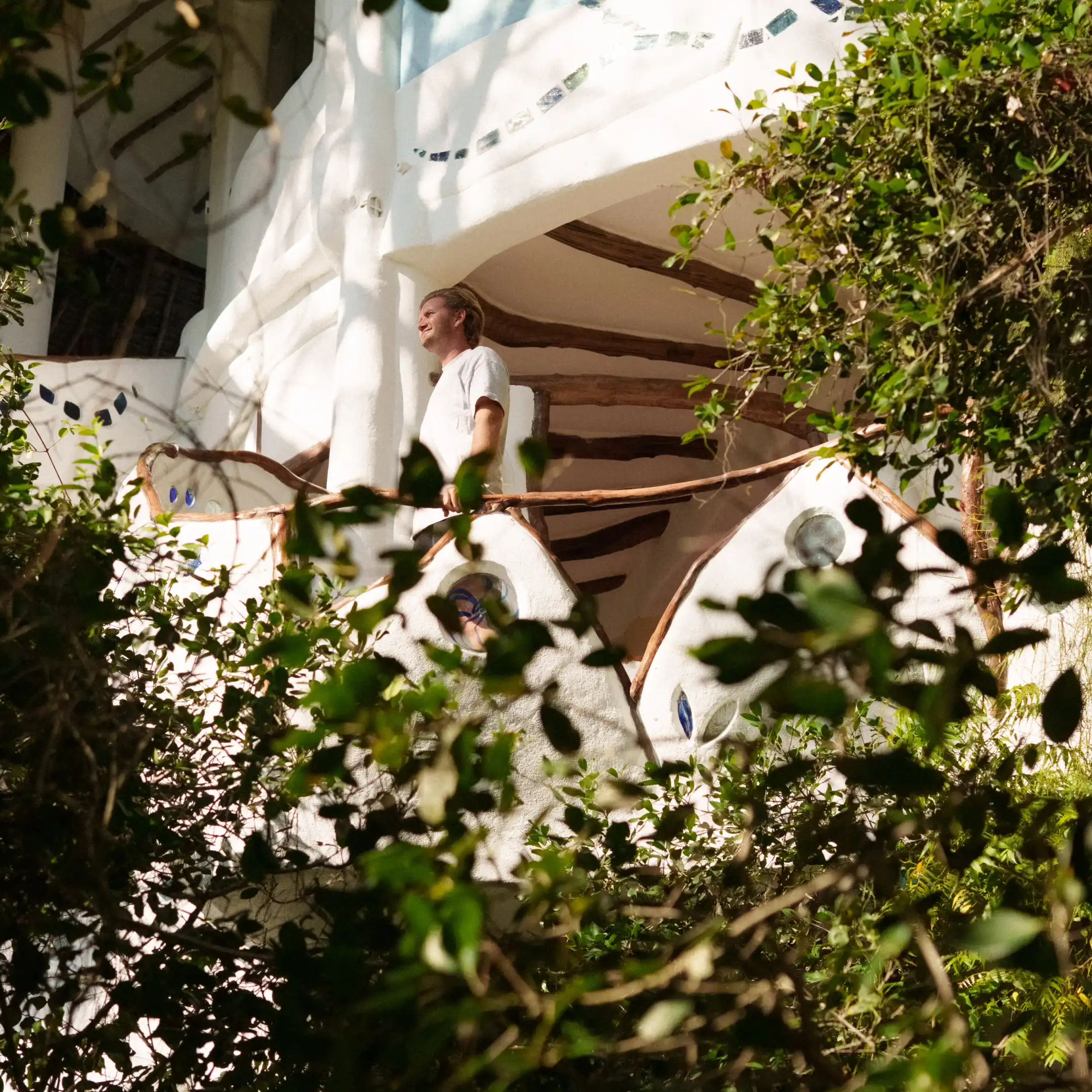 A man in a white shirt standing on a curved white balcony adorned with glass-mosaic inlays, framed by wooden beams and dense tropical foliage