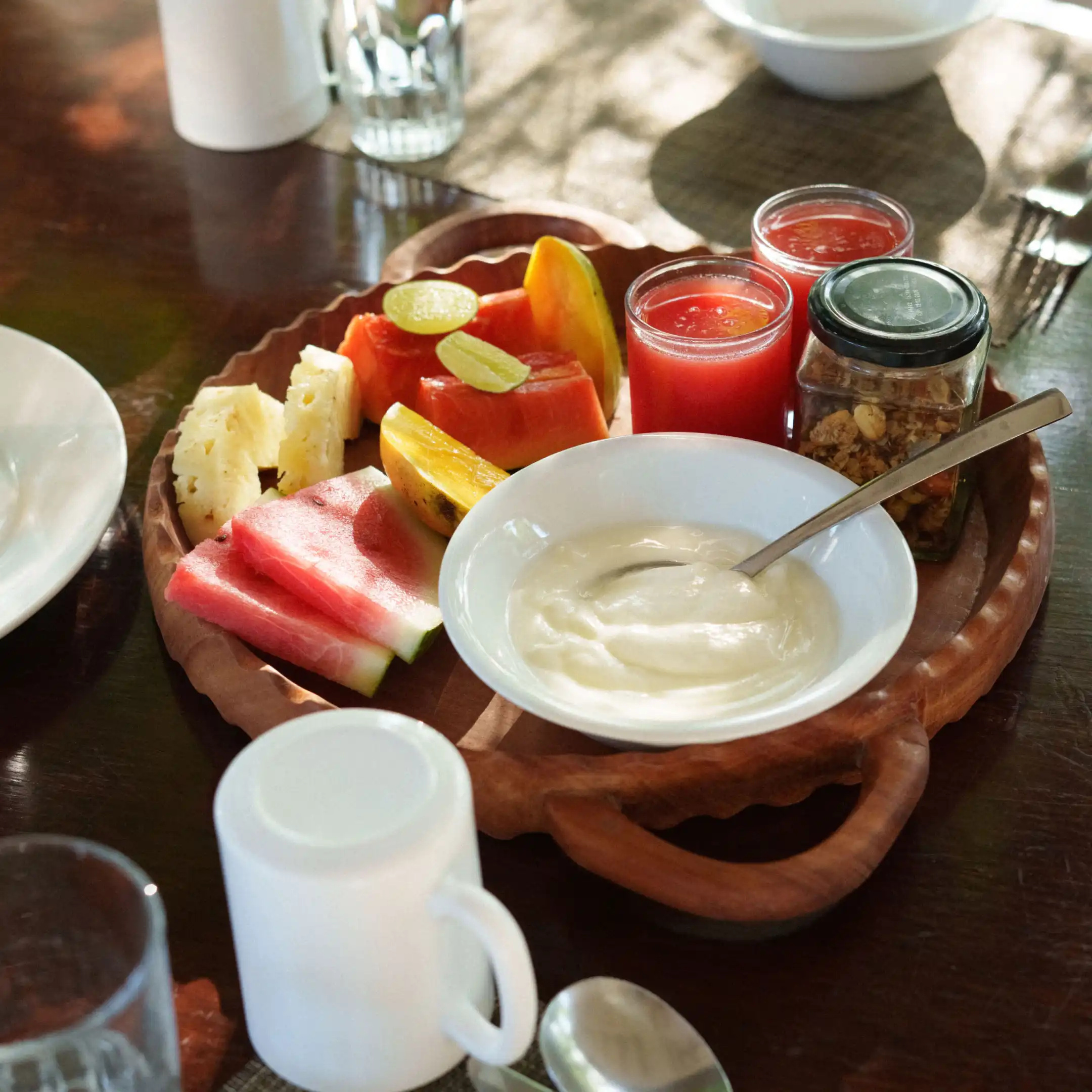 Wooden breakfast tray holding sliced watermelon, pineapple, papaya wedges, a bowl of yoghurt with spoon, a jar of granola, two glasses of red juice, and a white mug on a dark wood table