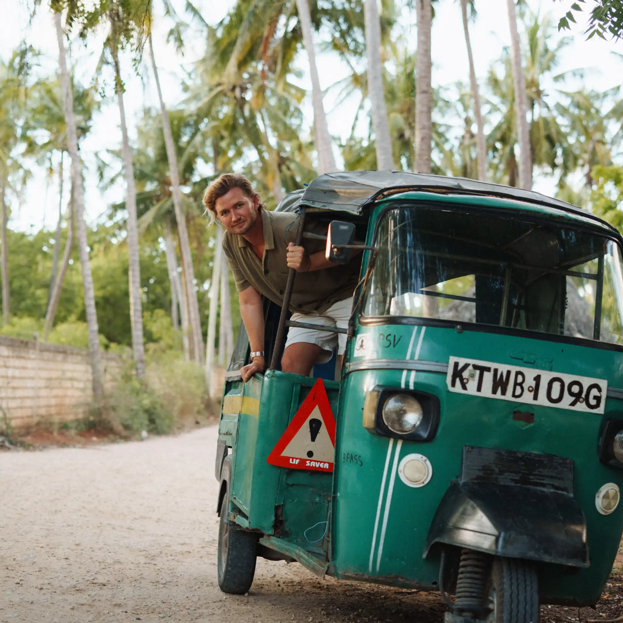 A young man leaning out of the open side of a green three-wheeled tuk-tuk, on a sandy road lined with tall coconut palms