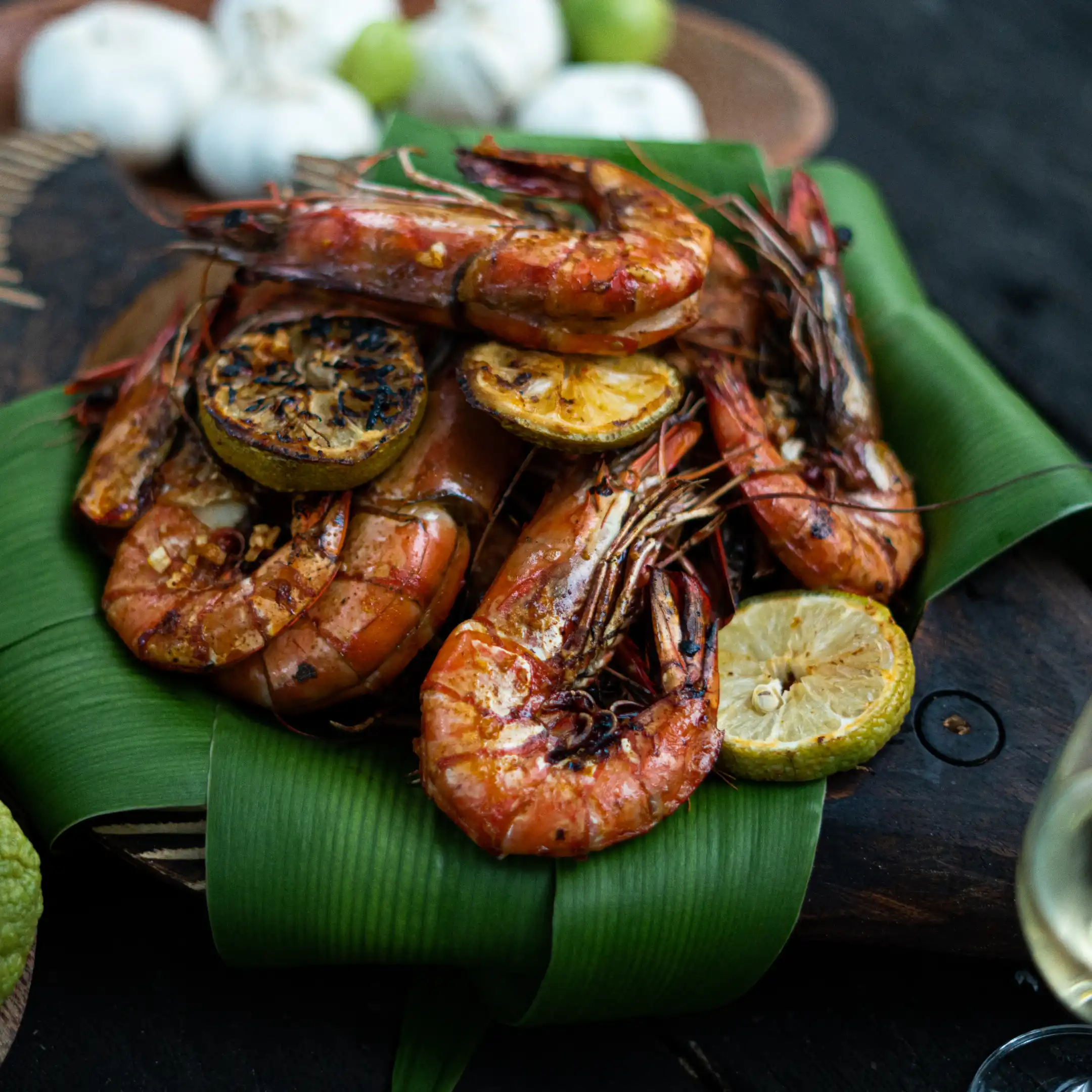 Charcoal-grilled prawns with glossy shells arranged on a banana leaf, garnished with seared lemon slices on a wooden serving board